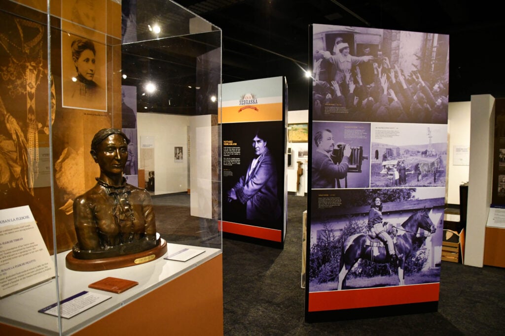Discover the Nebraska History Museum exhibit featuring a bronze bust of Susette LaFlesche Tibbles in a glass case, alongside informational panels displaying historical photos and text about notable figures and events.