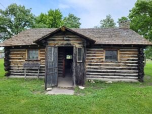 A small log cabin with a wooden shingle roof, front door open, two windows, and a sign reading “Log Cabin Built by WPA,” surrounded by grass and trees.