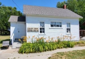 A small white wooden building with a sign reading "Valley Grove School Dist. 1873-1957," surrounded by orange flowers and green grass under a clear blue sky.
