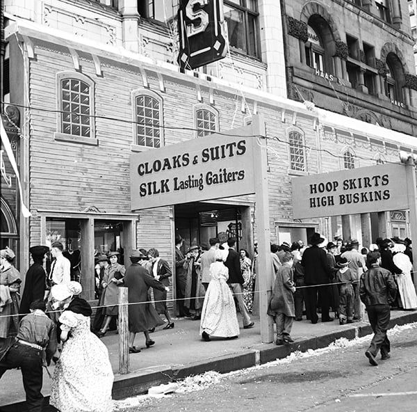 Crowd of people in period clothing walk past a wooden storefront advertising cloaks, suits, silk gaiters, hoop skirts, and high buskins, with historic buildings in the background.