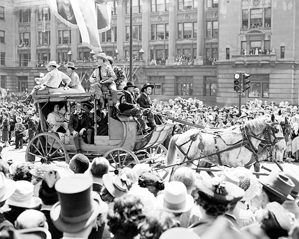 A horse-drawn stagecoach carrying people moves through a crowded city street during a parade, with onlookers in hats lining both sides.