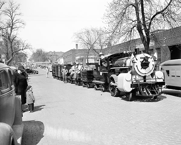 A small train with several passenger cars carries people down a brick-paved street lined with trees and buildings; bystanders watch from the sidewalk.