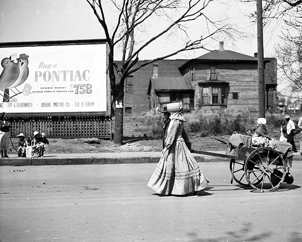 A woman in a long dress and bonnet walks beside a cart on a city street; a Pontiac billboard and old wooden house are in the background.