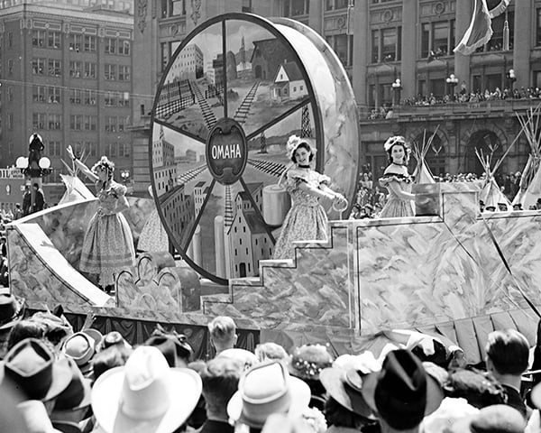 A parade float with a large "Omaha" wheel and three women in period costumes passes through a crowd of spectators wearing hats in a city setting.