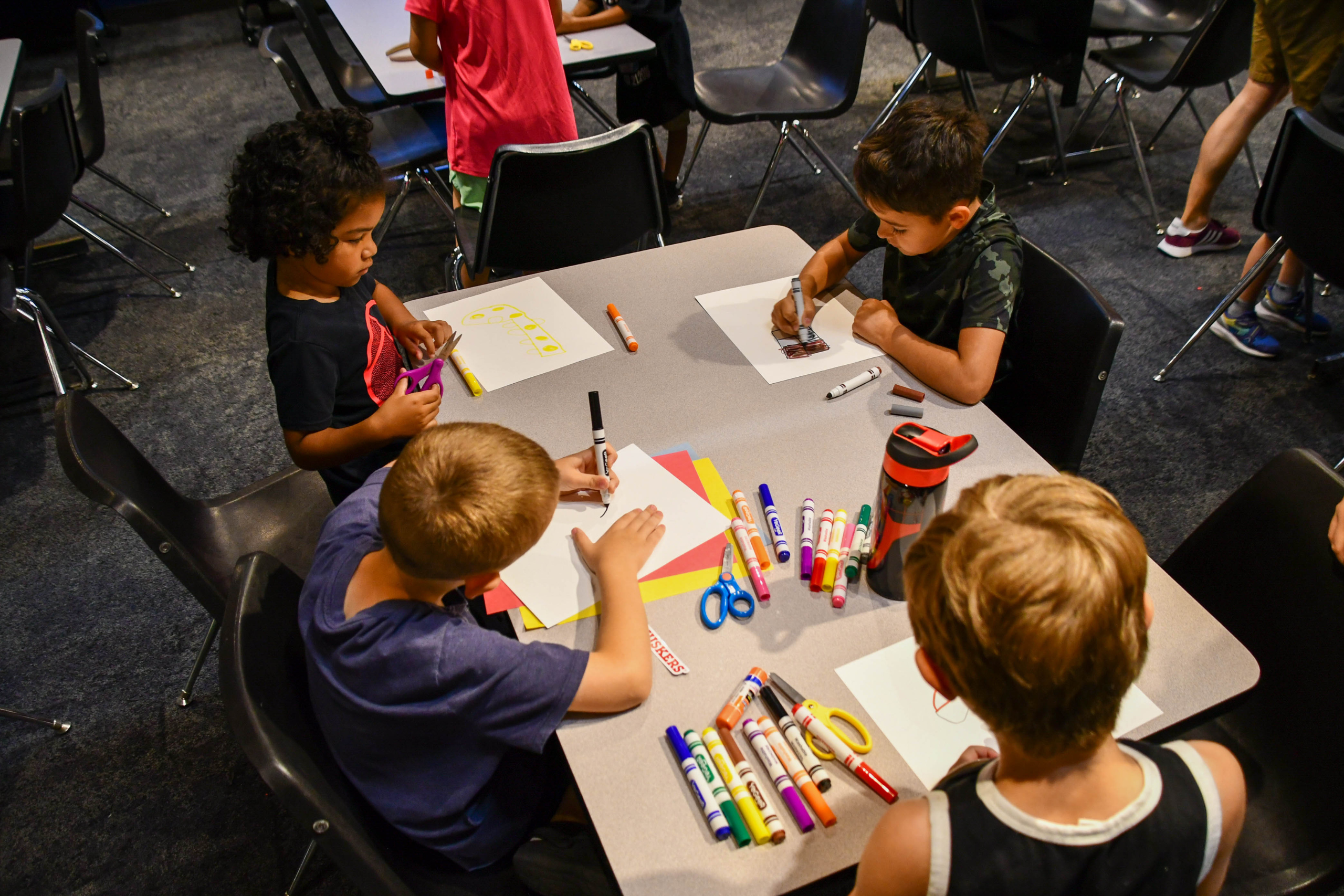 Four children sit at a table drawing on paper with markers and colored pencils, fully immersed in their Summer Workshops activities. Art supplies are spread across the tabletop; empty chairs surround them.