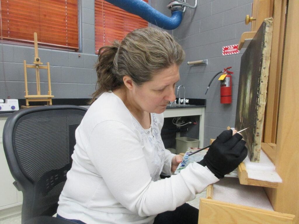 A woman wearing a white shirt sits at a desk carefully restoring a painting with a small brush in a workshop setting.
