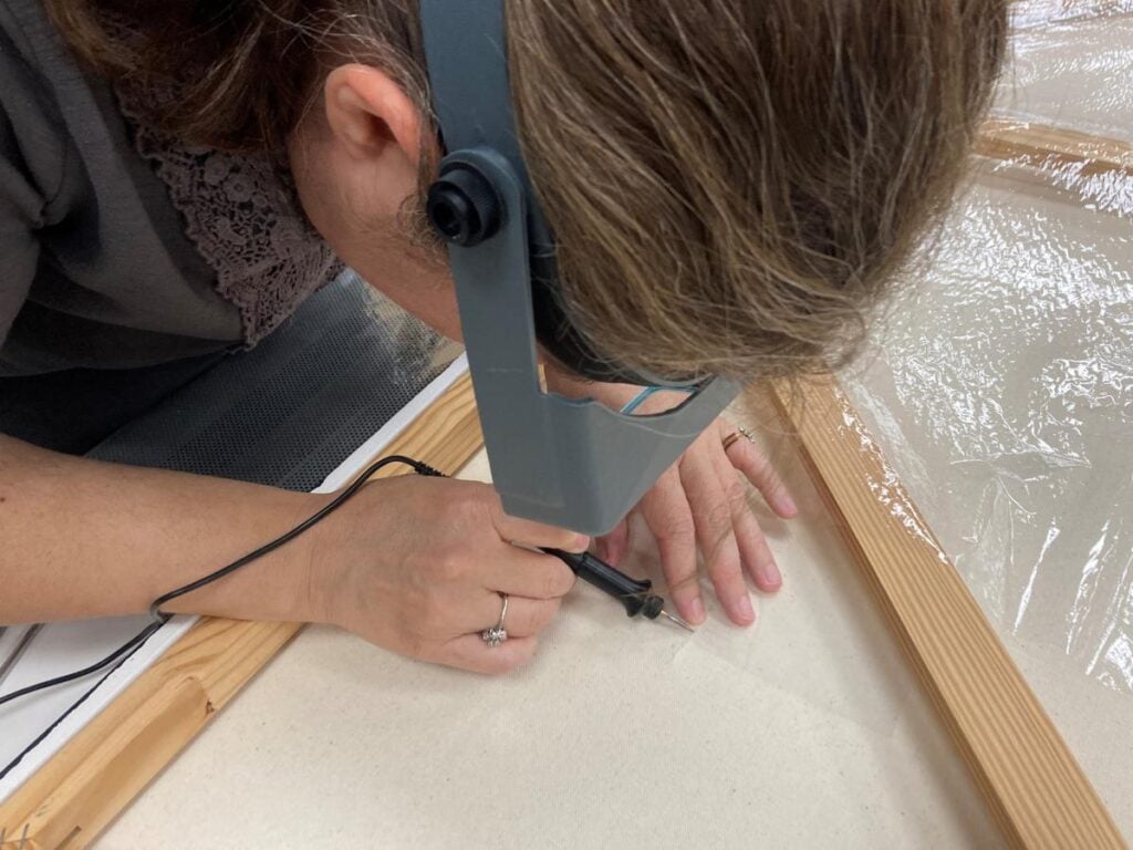 A person wearing magnifying glasses uses a handheld tool to work on the edge of a canvas stretched on a wooden frame.