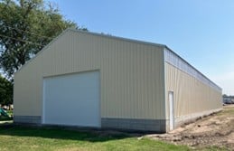A large beige metal storage building with a white garage door and a side entry door, situated on a grassy lot under a clear sky.