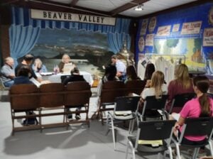 A group of people sits facing a panel at a table in front of a mural and signs labeled “Beaver Valley,” with vintage posters and garments displayed to the side.