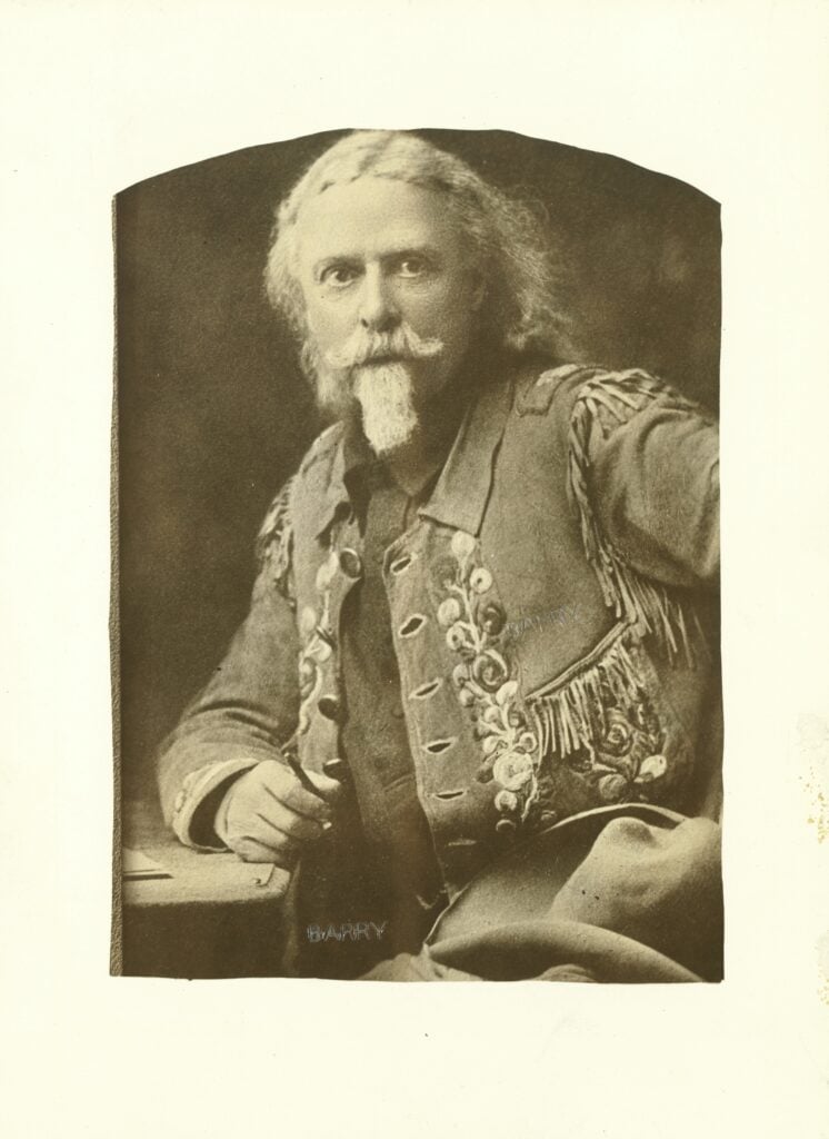 Sepia portrait of Buffalo Bill as older man with white hair and mustache, wearing a fringed, decorated jacket, seated and facing the camera.