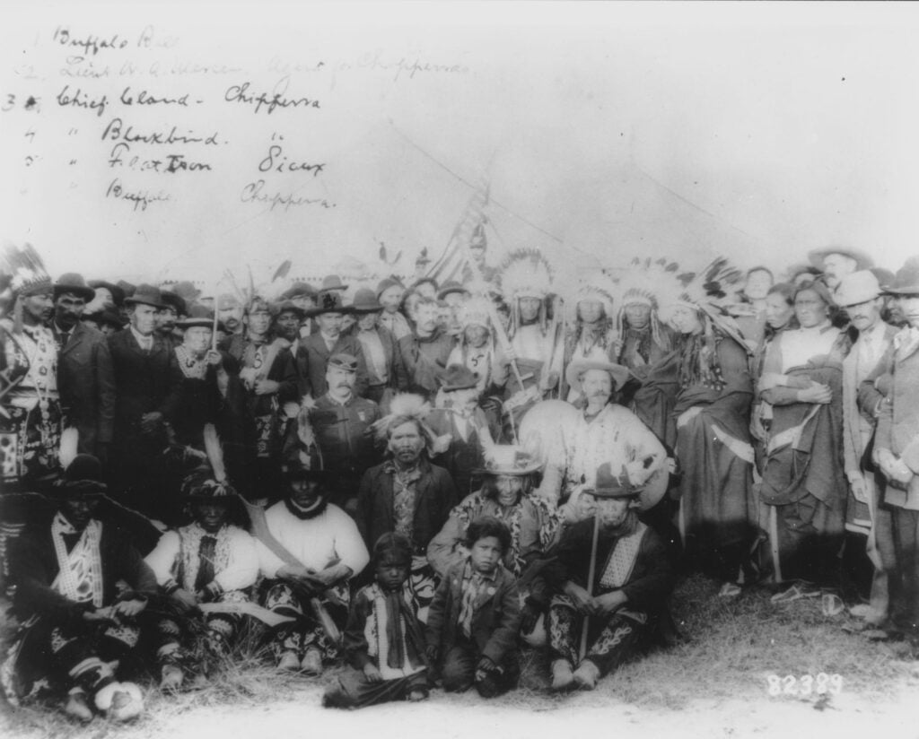 Large group of Native American men, women, and children in traditional clothing and headdresses pose outdoors with a few non-Native men; handwritten notes appear at the top of the image that list identified people.