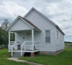 Small white wooden house with a front porch, steps, and railing, set on a grassy lawn under a partly cloudy sky.