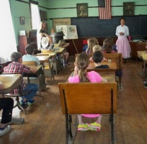 Children sit at desks in a classroom with wooden floors, while a woman stands at the front near a chalkboard and an American flag hangs above.