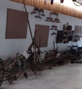 Antique farming tools and equipment displayed against a white wall inside a building with a wooden ceiling.