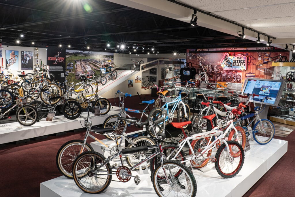 A well-lit indoor exhibit displays a variety of vintage BMX bikes on raised platforms, with informational signs and a mural in the background.