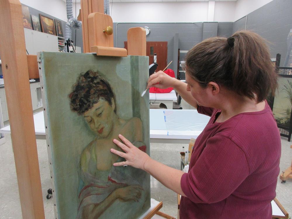 A woman in a red shirt restores a painting of a woman using a brush in an art studio.