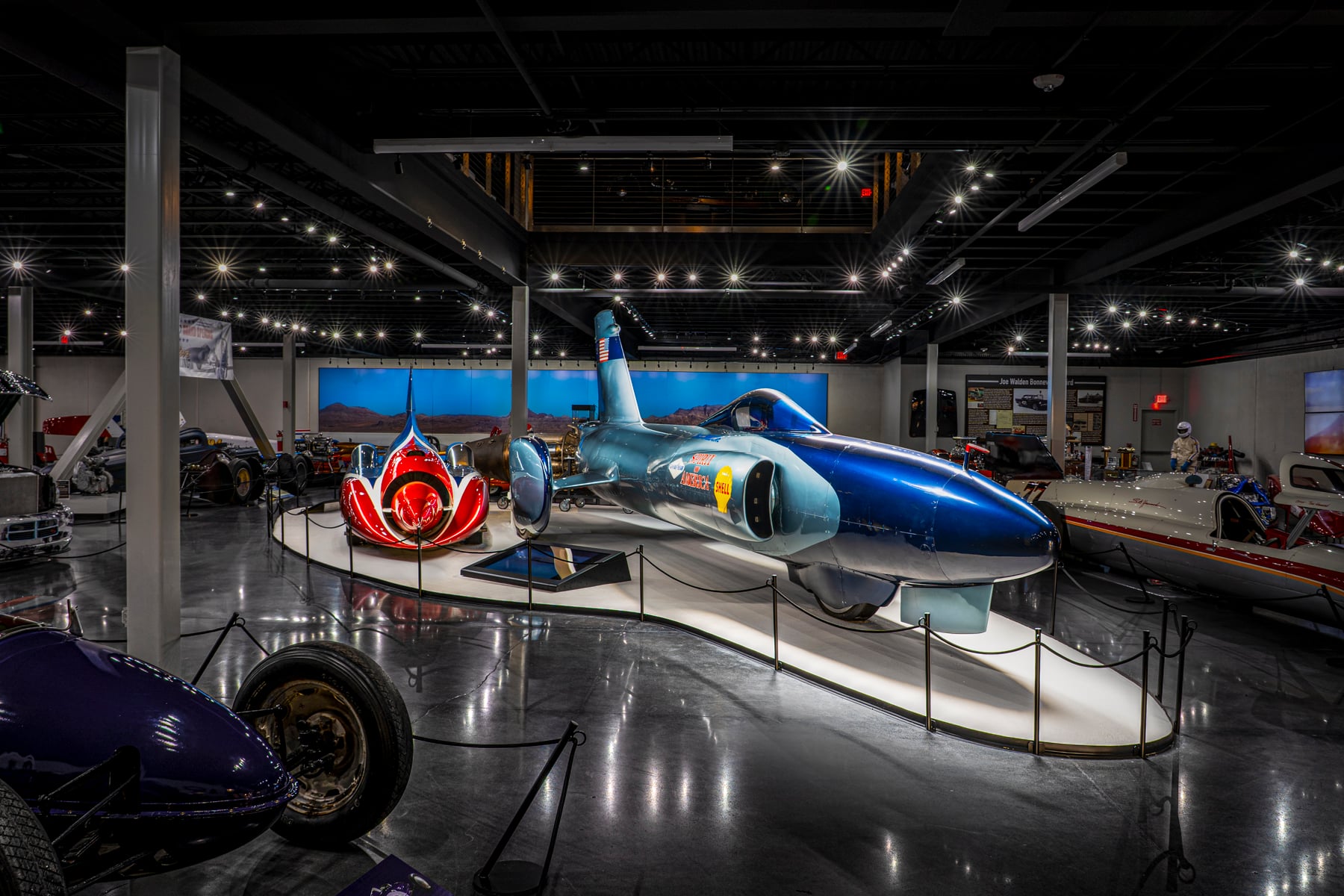 A blue land speed record car and a red jet engine are displayed in a museum surrounded by other vehicles, ropes, and bright overhead lights.