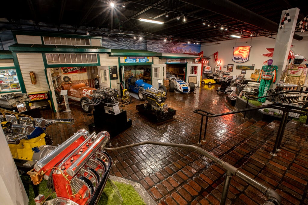 Indoor museum exhibit features vintage race cars, car engines, and memorabilia on brick flooring, with bright overhead lighting and retro signs on the walls.