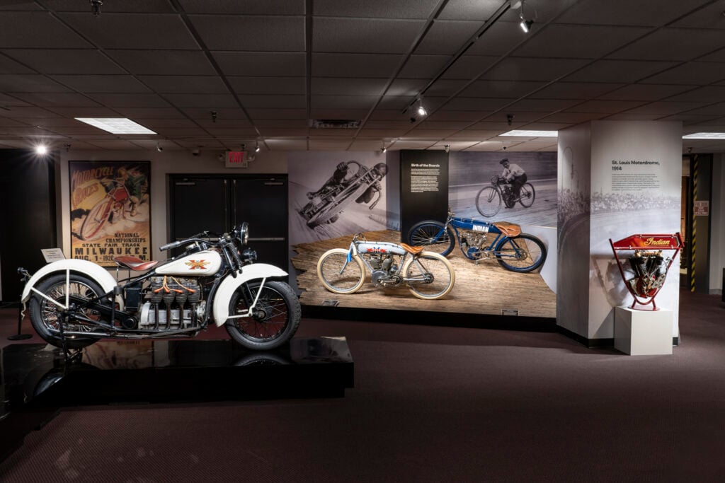 A museum exhibit displays vintage motorcycles and related memorabilia, including posters and photographs, against a background of brown carpet and white walls.