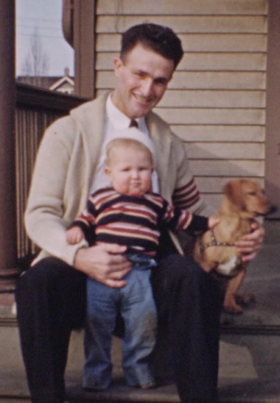 A man sitting on a porch steps holds a toddler on his lap, with a small dog sitting beside them.