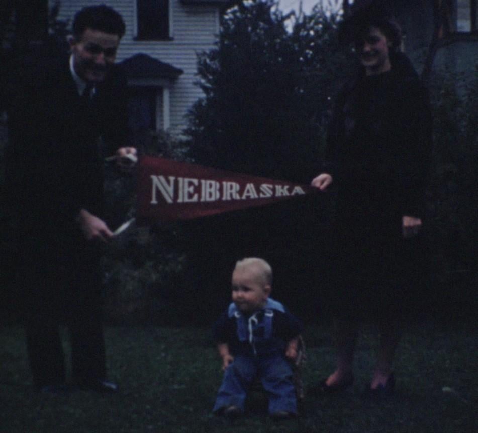 A man and woman standing outdoors holding a red Nebraska pennant over a small child sitting on the grass.