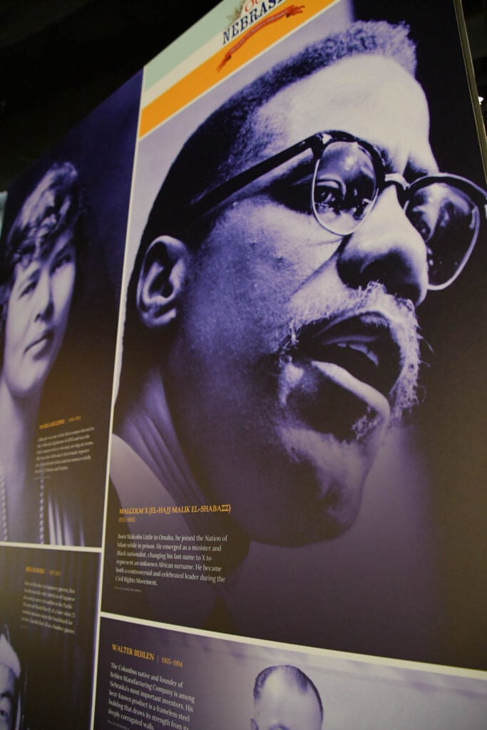 A display panel featuring a large portrait of Malcolm X, with biographical text below and smaller panels for other historical figures.